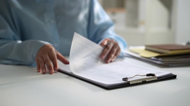 Person in blue shirt reviewing document and paperwork at office desk, turning pages on clipboard with pen and paper, carefully checking report in bright, softly blurred workspace with calm focus
