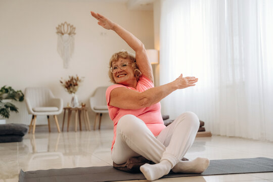 Smiling older woman stretching with raised arms while seated on a yoga mat in a bright modern living room, showcasing flexibility and wellness at home indoors