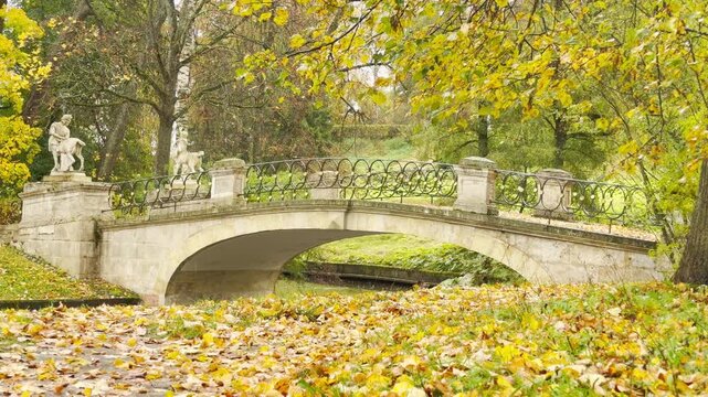 An old stone bridge with centaur statues stands as soft rain falls in slow motion. Golden leaves inspire peaceful nostalgia. This view is in Pavlovsk Park, Russia.