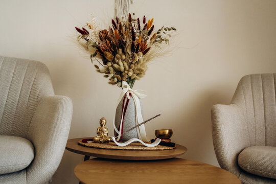 Dried botanical bouquet in ceramic vase with ribbons and small figurine between upholstered armchairs, styled on a wooden table in a neutral interior