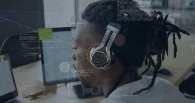 Working dreadlocked man wearing white shirt and headphones typing code at office desk with monitors