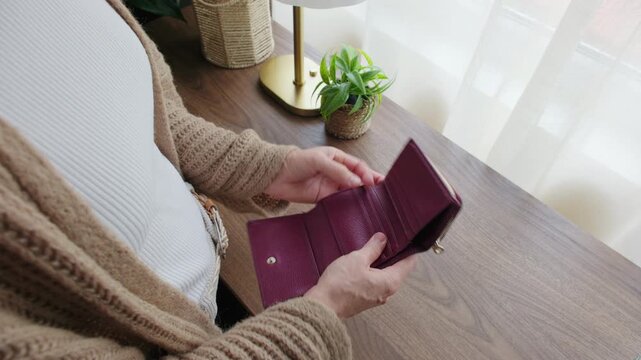 High-angle shot of a woman opening her wallet, finding no banknotes, and pouring out the last coins into her hand at home.