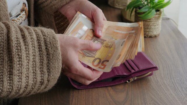 Close-up of female hands taking a thick stack of fifty euro banknotes from a leather wallet, counting them, and throwing the cash onto a wooden table.