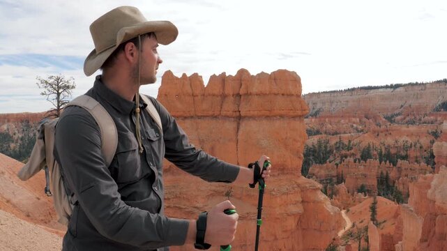 Caucasian male tourist wearing safari hat and backpack, leaning on trekking poles while admiring majestic red rock hoodoos at bryce canyon national park during sunny day
