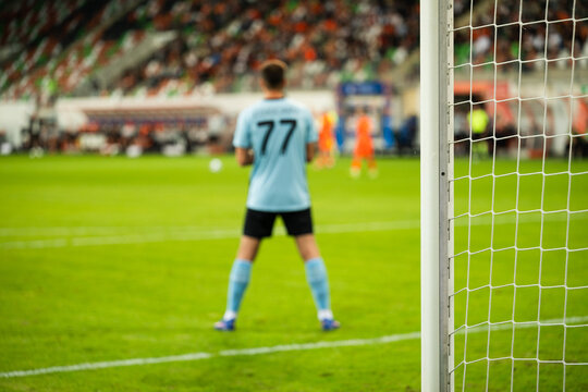 Football goalkeeper wearing number 77 watches the action from inside the goal during a match, with the net in the foreground and players in the distance.