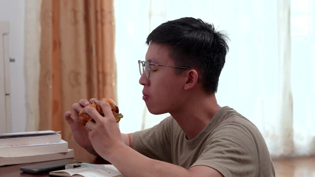 young Asian male student or freelancer taking a bite of a burger and licking his fingers enthusiastically at his desk