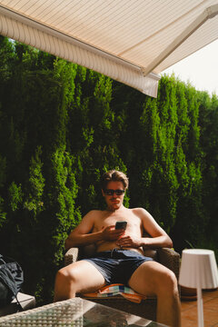 Shirtless young man using smart phone while sitting on chair under awning shade