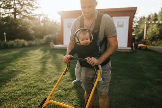 Father mowing lawn while carrying male toddler wearing noise canceling headphones in front carrier