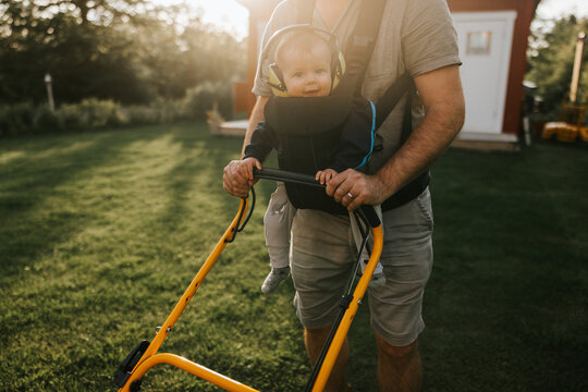Smiling baby riding in front facing carrier while father pushes yellow lawn mower handle