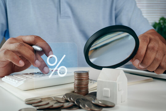 Business professional using calculator and magnifying glass near house model and coins with percentage icon overlay, concept of mortgage interest rate, house loan, and property investment.