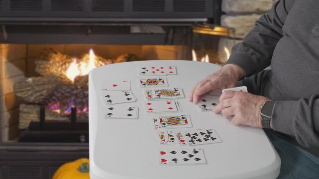 A person arranges playing cards for a game of solitaire at a table beside a lit fireplace indoors.