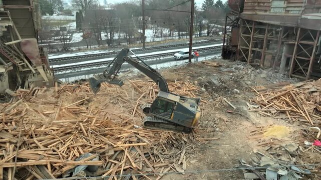 An excavator moves through piles of timber debris at a demolition site beside railway tracks, with snow on the ground and bare trees in the background.