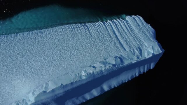 A near-vertical drone view slowly pulls back from a huge white-blue iceberg in Antarctic waters. Parallel grooves, sculpted ice texture and sparkling sea emerge in the widening overhead composition.