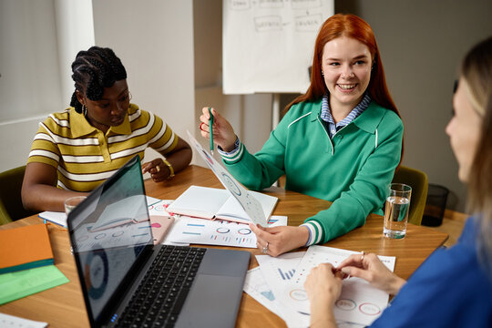 Diverse young adult Caucasian and African American women analyzing financial charts during business meeting in coworking office. Useful for corporate marketing and teamwork campaigns