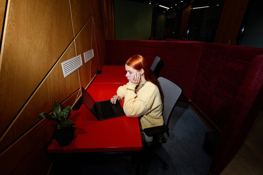 High angle shot of Caucasian young adult woman working on laptop in coworking office cubicle. Image suitable for remote work, freelance technology, corporate productivity themes