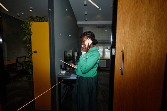 Young adult African American woman talking on smartphone while holding digital tablet in modern coworking office. Visual suitable for corporate communication and remote work marketing