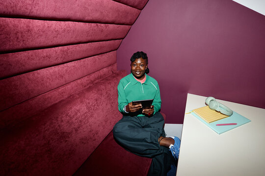 Portrait of young African American woman smiling while holding digital tablet in modern coworking office booth. Ideal for corporate branding, mobile technology, and remote work marketing