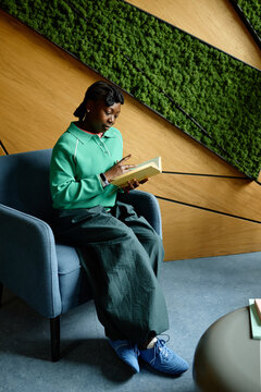 Vertical shot of young adult African American Black woman reading book in modern coworking office lounge. Ideal for corporate learning, professional development and workplace wellness campaigns