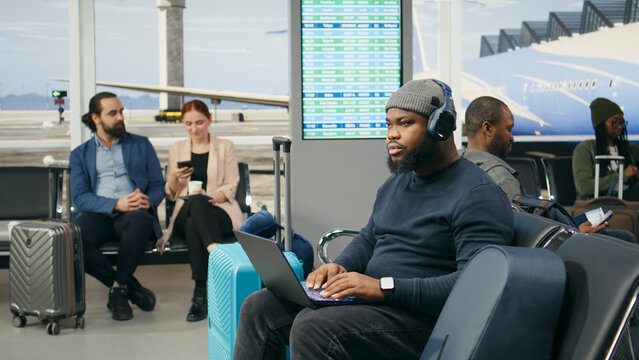 Airport traveler, laptop and remote work as man in knit hat types beside blue suitcase while waiting for flight. Productivity, business mobility and flexible working support tasks between journeys.