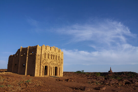Decorated mud mosque standing on rocky ground with a minaret in the distance under a blue sky, Sahel Region, Bani, Burkina Faso