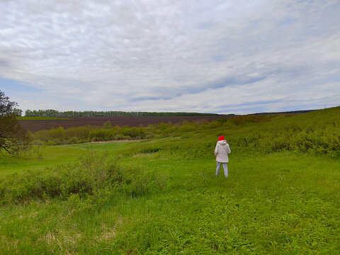 A woman in light clothing stands alone in a wide green meadow beneath patterned clouds, suggesting a quiet pause away from everyday routines