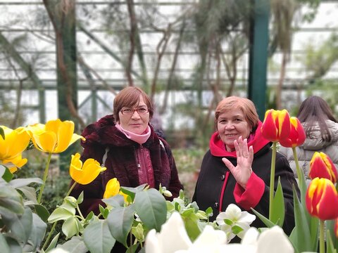 Two older women smile behind bright tulips, with one raising a hand toward the camera during a relaxed greenhouse outing