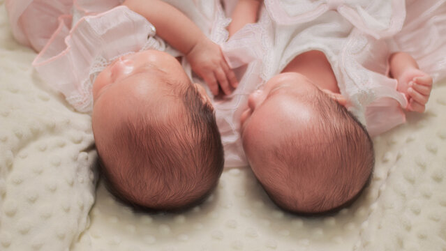 Newborn twin babies resting side by side on a soft cream blanket, sibling bonding, infancy, and newborn care. Wearing delicate white outfits, capturing a tender family moment.