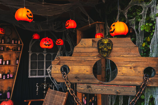 Spooky Halloween decoration with wooden pillory, jack o lantern pumpkin lanterns, golden skull, spider webs, and eerie potion shelves. Halloween haunted house background with stocks pillory device.