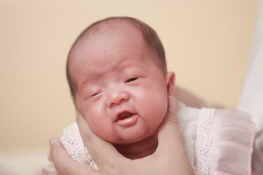 Infant care concepts, newborn, cradle the head, tenderness, and early family bonding. Close up portrait newborn baby gently supported adult hand, wearing white outfit against warm pastel background.