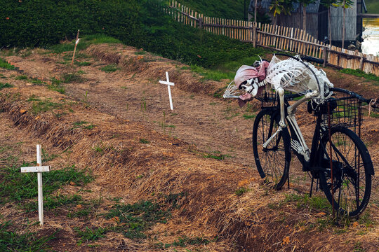 Skeleton prop riding vintage bicycle with broomstick in lake park graveyard. Skeleton prop slumped on bicycle wrapped in spider webs for Halloween outdoor display. Halloween outdoor decorations.