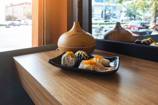 Sushi dish on window counter bar with ultrasonic humidifier. Assorted Japanese sushi, including nigiri and gunkan topped with roe next to wood grain aroma diffuser oil diffuser by window view.