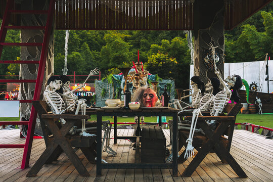Halloween Dinner Party with Skeletons and Severed Head Prop. Human skeletons seated on wooden bench at table, mimicking dinner party. Idea concept for Halloween props, pirate theme party decorations.