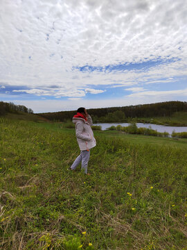 A woman pauses near a pond in a grassy landscape, with a dramatic cloud pattern and open space emphasizing a reflective countryside escape