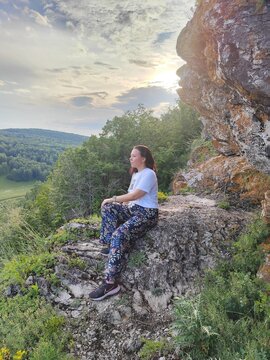 A woman relaxes on a stone outcrop beneath a softly lit sky, blending an informal travel portrait with a broad countryside view