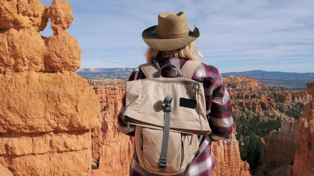 Female hiker wearing a backpack and hat standing on cliff edge, admiring red rock hoodoos of bryce canyon national park during an outdoor adventure. Camera panning around standing hiker on top canyon
