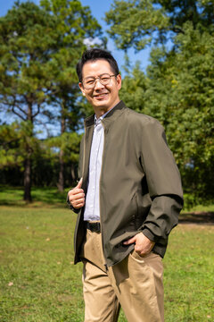 A confident, mature Chinese man, dressed in business-casual attire, stands outdoors against a backdrop of trees and a blue sky.