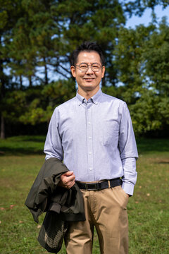 Portrait of smiling middle-aged Asian man with glasses, holding jacket, wearing light blue button shirt in park