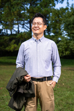 Portrait of smiling middle-aged Asian man with glasses, holding jacket, wearing light blue button shirt in park