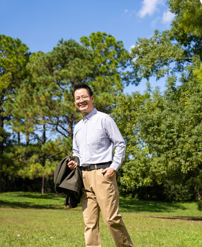 Portrait of smiling middle-aged Asian man with glasses, holding jacket, wearing light blue button shirt in park