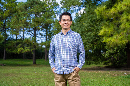 Portrait of smiling middle-aged Asian man with glasses in blue plaid shirt standing in park on sunny day