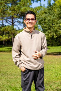 A middle-aged Asian man wearing glasses stands in a park.