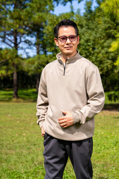 A middle-aged Asian man wearing glasses stands in a park.