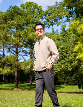 A middle-aged Asian man wearing glasses stands in a park.