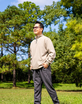A middle-aged Asian man wearing glasses stands in a park.