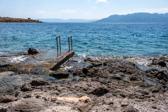 Clear turquoise waters lap against the rugged rocky shore near Perdika village on Aegina Island, offering a scenic view of Moni Island and distant sailboats in the Saronic Gulf