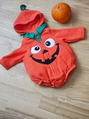 Orange Halloween Costume with Pumpkin Beside It on Wooden Floor,Bright orange fleece Halloween costume featuring a funny jack-o'-lantern face, large cartoon eyes, a black smiling mouth © Алеся Макаревич