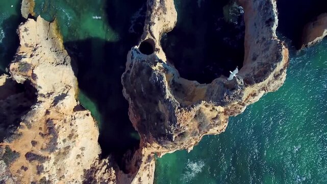 Drone camera flying directly above rugged sandstone sea arches and turquoise atlantic waters along praia dos tres irmaos in algarve portugal, revealing birds gliding over the coast