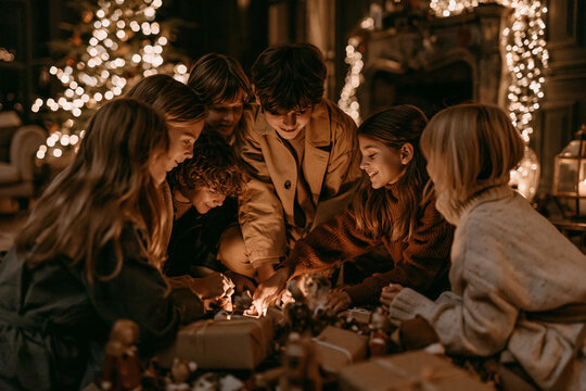 A group of six children gathered in a cozy dimly lit room huddled together over Christmas gifts near a glowing fireplace and tree sharing a warm magical holiday moment of joy