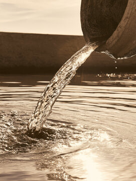 Concrete outfall pipe releasing clear water into retention basin under warm morning sunlight, creating ripples, splashes, reflections, and calm industrial atmosphere
