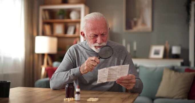 Senior man uses a magnifying glass to read a medicine leaflet at table.